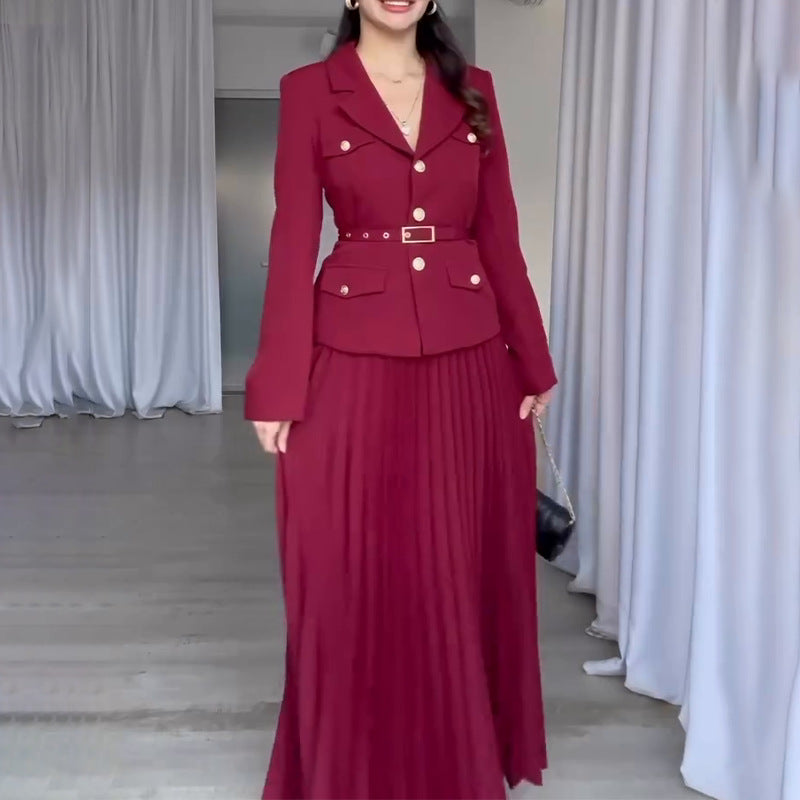 Elegante Frau in rotem Blazer und plissiertem Maxirock, posiert in modernem Raum mit weißen Vorhängen. Modetrend, Damenmode, stilvolles Outfit.