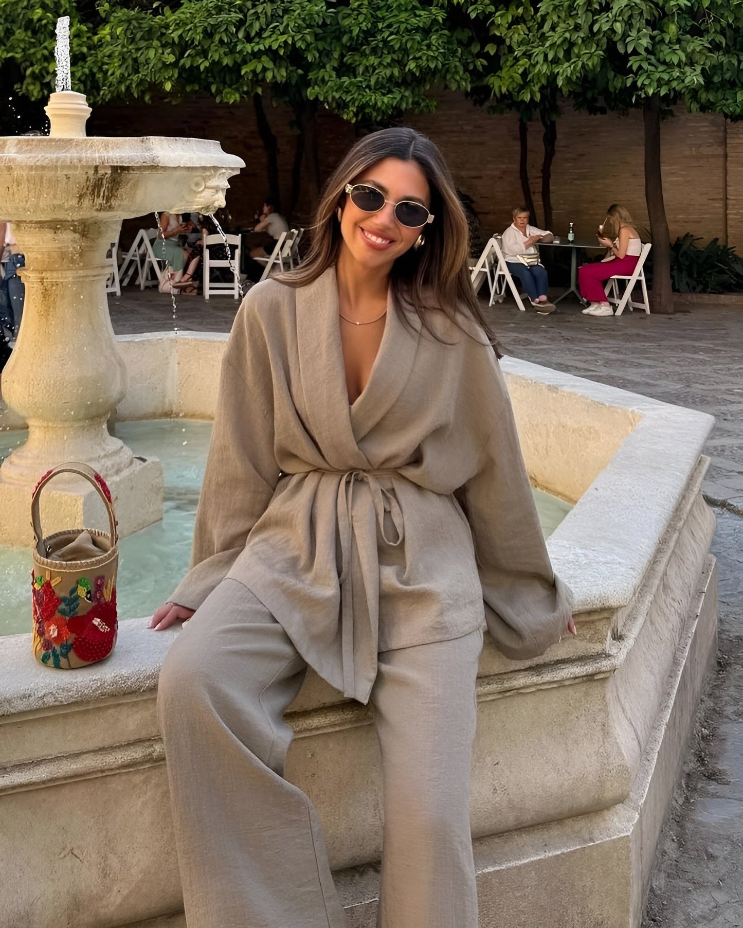 Eine Frau in beiger Leinenkleidung sitzt lächelnd auf einem Brunnenrand. Sie trägt Sonnenbrillen, im Hintergrund sind Bäume und Menschen an Tischen.