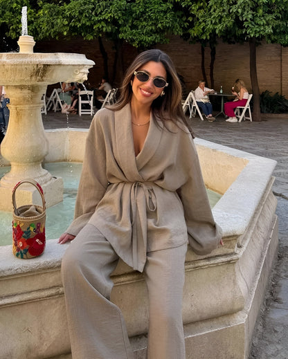 Eine Frau in beiger Leinenkleidung sitzt lächelnd auf einem Brunnenrand. Sie trägt Sonnenbrillen, im Hintergrund sind Bäume und Menschen an Tischen.