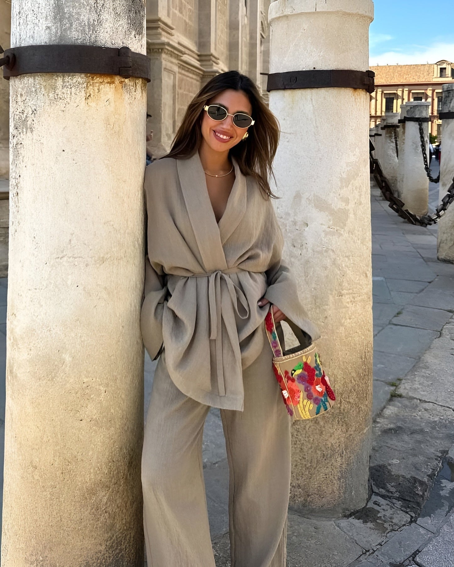 Frau in beigem Outfit lehnt an Säule, trägt Sonnenbrille und bunte Tasche. Stilvolle Mode, lässiger Look, urbane Kulisse.