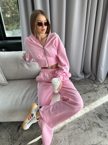 Frau in rosa Jogginganzug mit Sonnenbrille sitzt auf Sofa, hält Kaffeebecher. Modetrend, Freizeitkleidung, gemütlicher Stil.