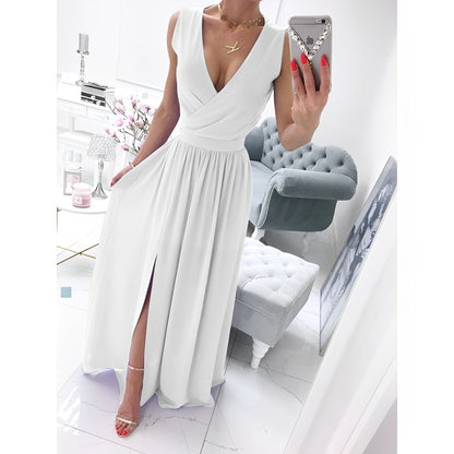 Frau in elegantem weißen Maxikleid mit V-Ausschnitt und Schlitz, steht in modernem Wohnzimmer mit grauem Sessel und Spiegel, hält Smartphone.
