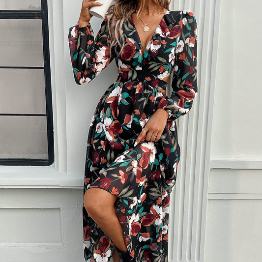 Frau in langem, geblümtem Kleid vor weißer Wand. Elegantes Design, florales Muster, Herbstmode, stilvolle Damenbekleidung, Fenster im Hintergrund.