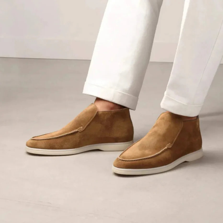 Braune Wildleder-Stiefeletten für Herren auf hellem Boden, kombiniert mit weißen Hosen. Elegante, moderne Herrenschuhe, ideal für stilvolle Outfits.