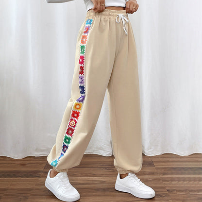 Beige Jogginghose mit buntem Blumenmuster an den Seiten, elastischem Bund und weißen Schnürsenkeln, kombiniert mit weißen Sneakers auf Holzboden.