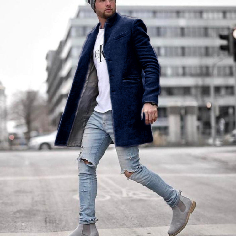 Mann in blauem Mantel und zerrissenen Jeans überquert Straße, moderne urbane Mode, graue Stiefel, Stadtgebäude im Hintergrund.