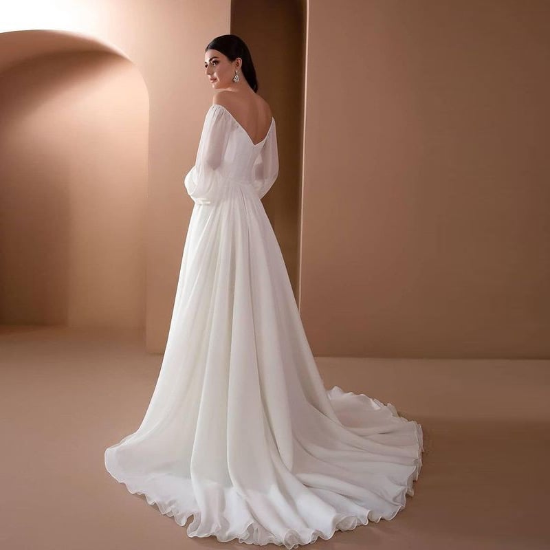 Braut in elegantem, langem weißen Hochzeitskleid mit tiefem Rückenausschnitt und fließendem Rock, steht vor minimalistischem, beigem Hintergrund.