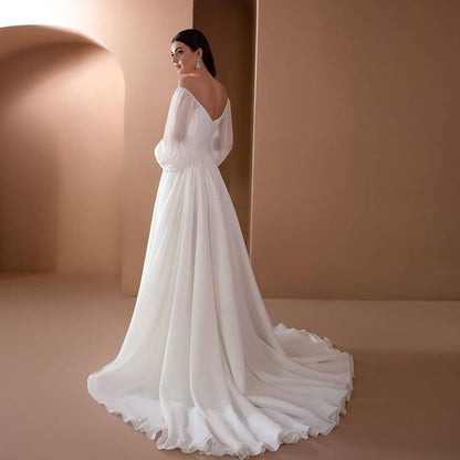 Braut in elegantem, langem weißen Hochzeitskleid mit tiefem Rückenausschnitt und fließendem Rock, steht vor minimalistischem, beigem Hintergrund.