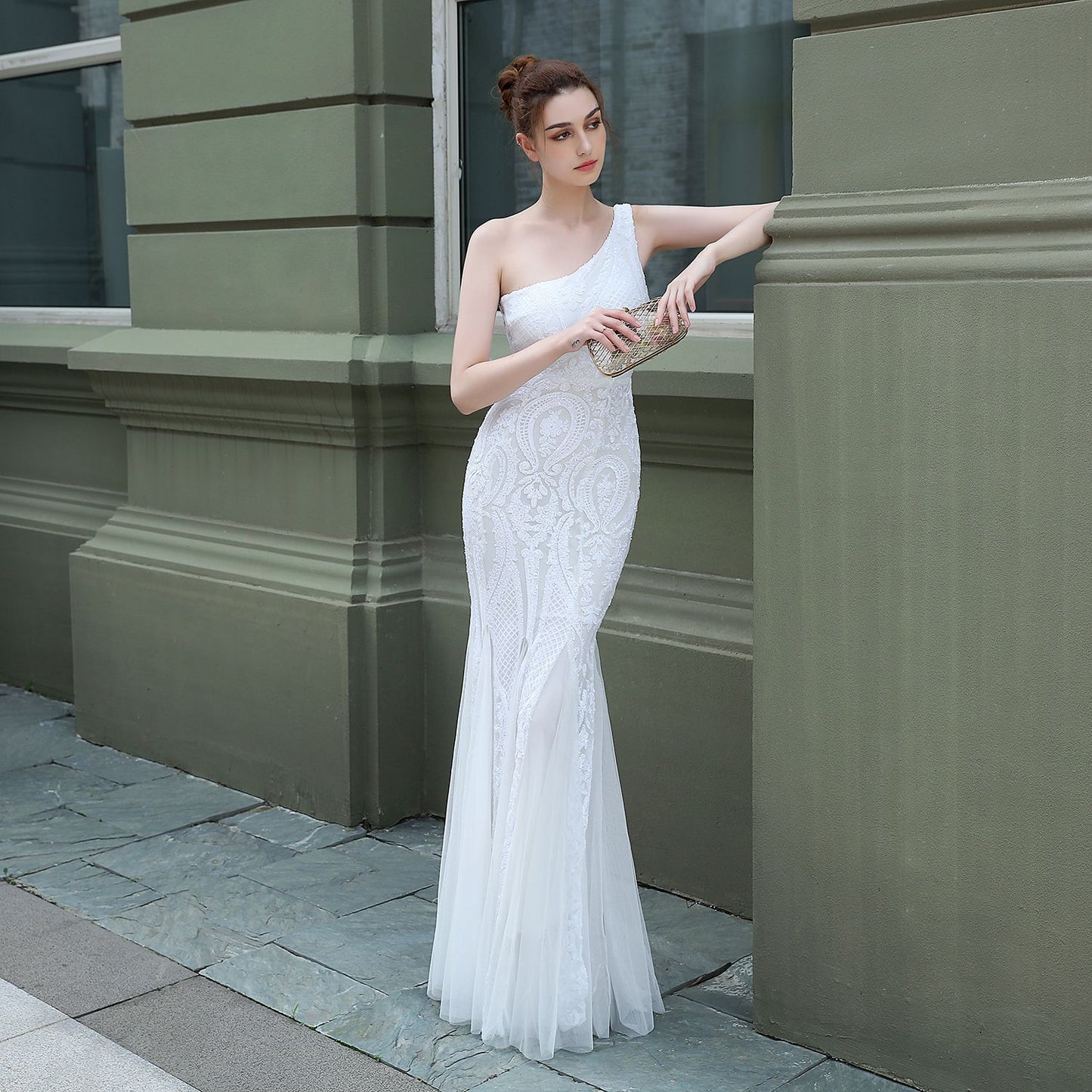 Frau in elegantem, einarmigem weißen Abendkleid mit Spitzenmuster lehnt an grüner Wand, hält eine glitzernde Clutch. Perfekt für Hochzeiten oder Galas.