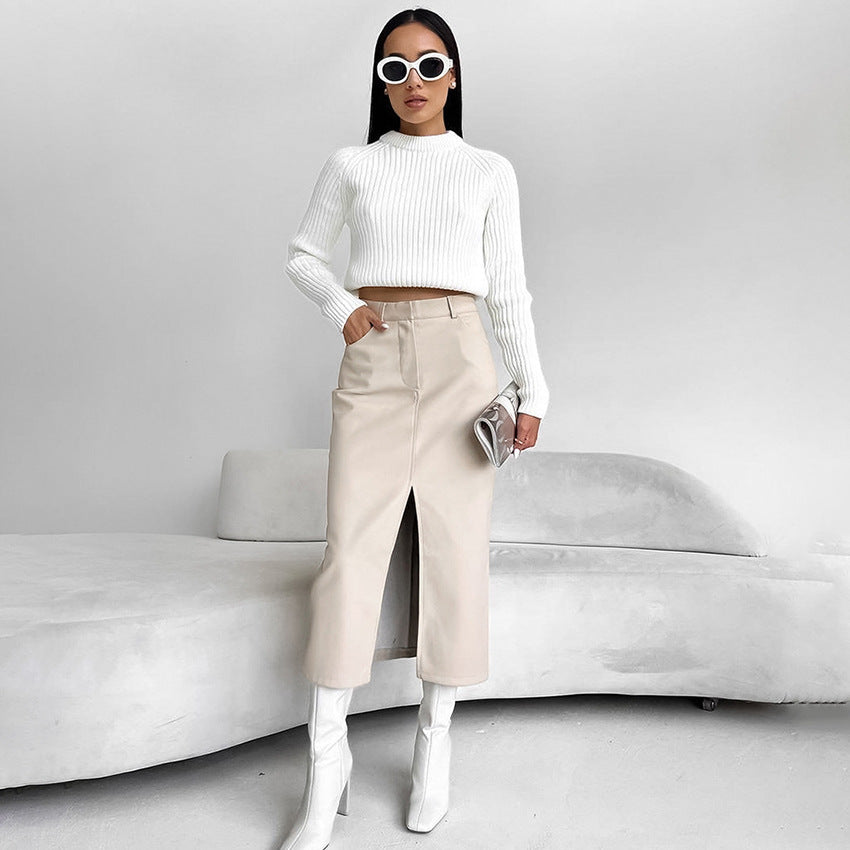 Frau in weißem Pullover und beigem Rock mit Schlitz, trägt weiße Stiefel und Sonnenbrille, steht vor grauem Sofa. Elegante Mode, minimalistischer Stil.