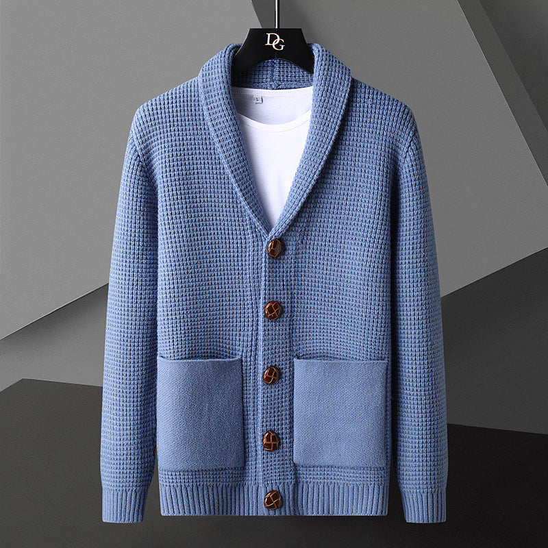 Blaue Strickjacke mit Schalkragen, Holzknöpfen und aufgesetzten Taschen, hängt auf einem Kleiderbügel vor grauem Hintergrund.