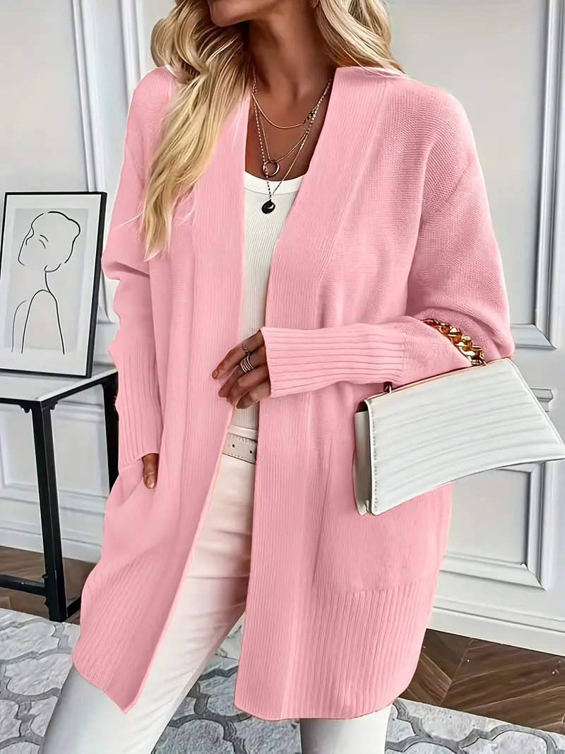 Frau in rosa Strickjacke, weißem Top und Hose, hält weiße Handtasche. Elegante Mode, lässiger Stil, modernes Outfit, Innenraum.