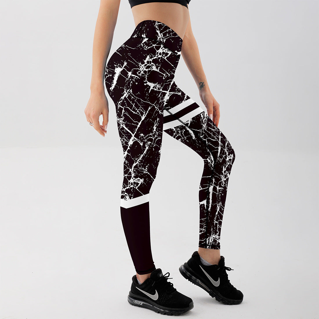 Frau in schwarz-weiß gemusterten Leggings und schwarzen Nike-Sneakern, seitlich stehend vor grauem Hintergrund. Sportmode, Fitness, Damenbekleidung.