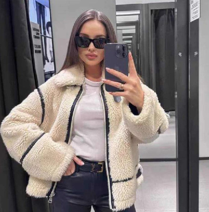 Frau mit Sonnenbrille macht Selfie in Umkleidekabine, trägt weiße Teddyjacke und schwarze Lederhose. Modetrend, Wintermode, Selfie-Stil.