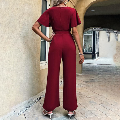 Frau in rotem Jumpsuit von hinten, steht auf gepflastertem Weg, trägt weiße High Heels. Elegante Mode, Sommeroutfit, stilvolle Damenbekleidung.