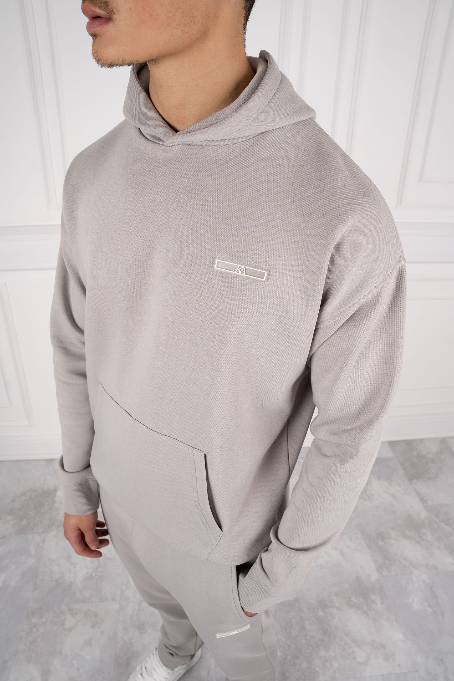 Mann in grauem Hoodie und Jogginghose, modernes Design, minimalistische Mode, Freizeitkleidung, weiße Wand im Hintergrund.