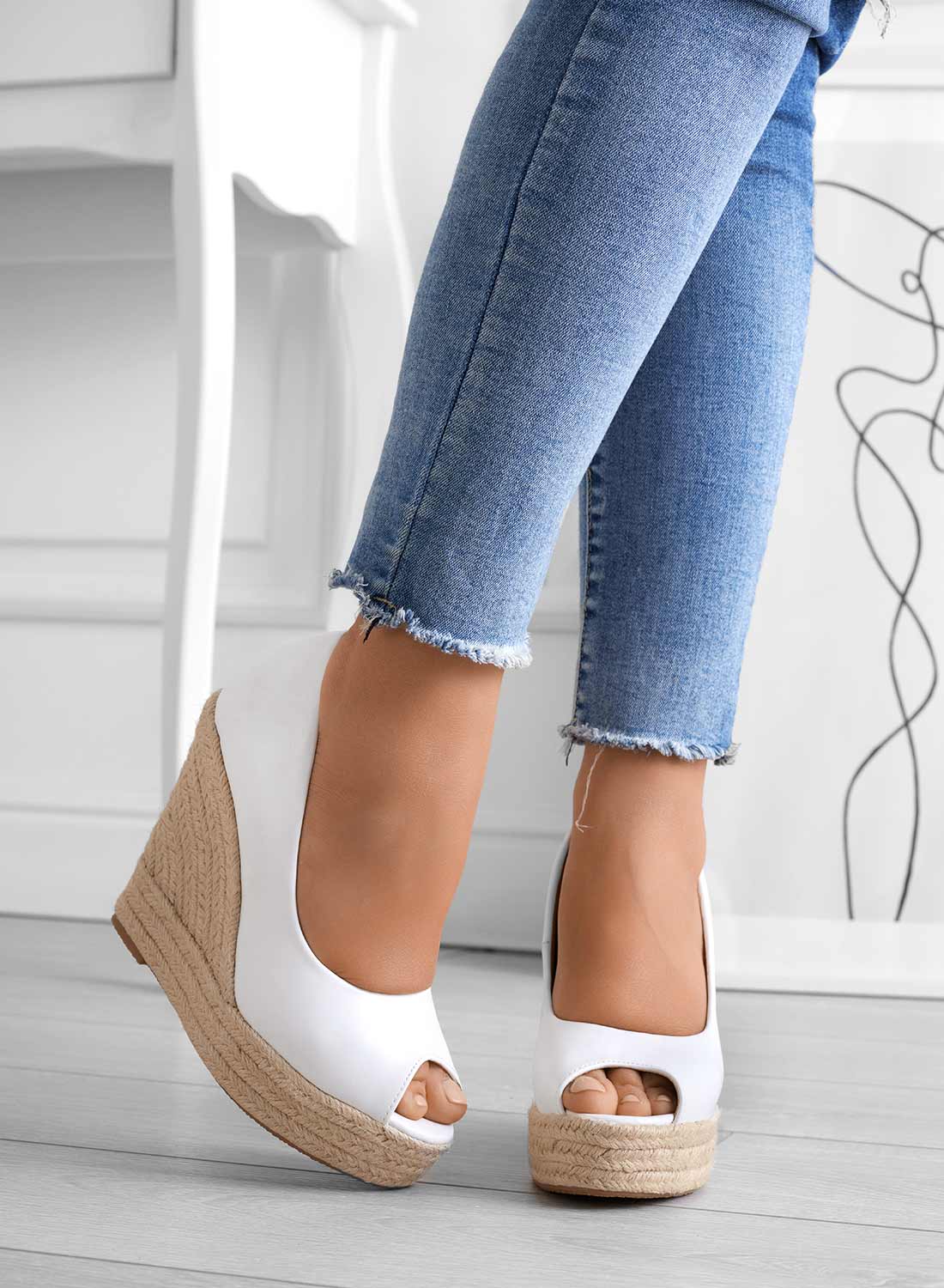 Frau trägt weiße Keilsandaletten mit Bastsohle und offener Spitze, kombiniert mit blauen Jeans. Modische Sommerschuhe, Damenmode, Casual-Look.