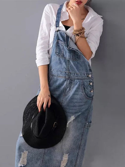 Frau in zerrissener Jeans-Latzhose und weißem Hemd, hält schwarzen Hut. Modetrend, lässiger Stil, Denim-Outfit, urbaner Look.