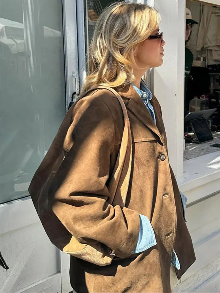 Blonde Frau mit Sonnenbrille trägt braune Wildlederjacke und Jeanshemd, steht vor einem Fenster. Modetrend, Herbstmode, Streetstyle.