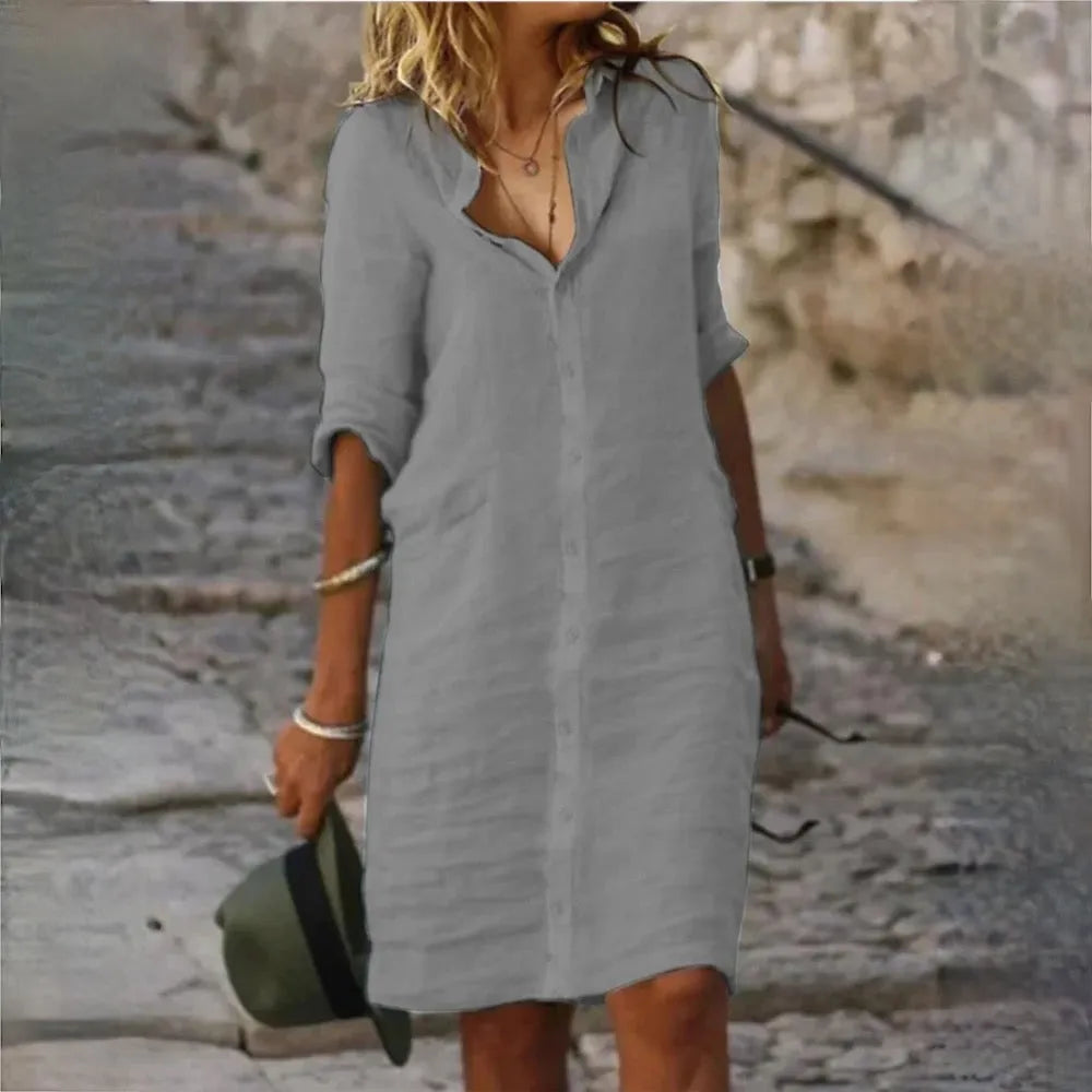 Frau in grauem Leinenkleid auf Kopfsteinpflaster, hält Hut in der Hand. Sommermode, lässiger Stil, elegantes Freizeitoutfit.