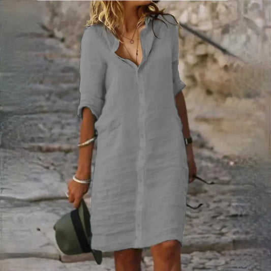 Frau in grauem Hemdkleid auf Kopfsteinpflaster, hält Hut in der Hand. Sommermode, lässiger Stil, Outdoor-Spaziergang.
