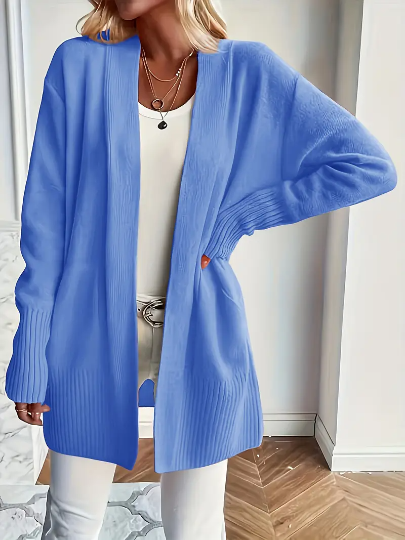 Frau in blauem Strick-Cardigan, weißem Top und Hose, steht in modernem Raum. Modetrend, Damenmode, lässiger Stil, Herbstoutfit.
