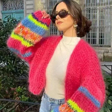 Frau in pinker, flauschiger Strickjacke mit bunten Streifen, Sonnenbrille und Jeans vor urbanem Hintergrund. Modetrend, Herbstmode, Streetstyle.