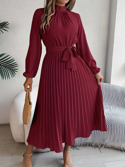 Frau in elegantem, langem, weinrotem Kleid mit Gürtel und Plissee, hält eine Strohtasche. Perfekt für Herbstmode und stilvolle Anlässe.