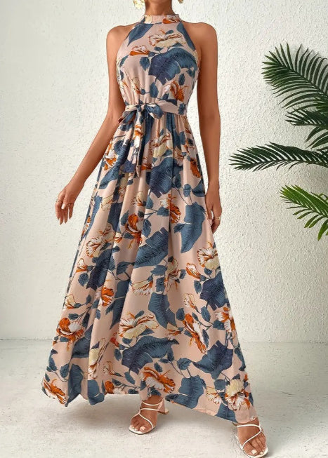 Frau in langem, ärmellosem Maxikleid mit tropischem Blumenmuster in Blau und Orange, steht vor weißer Wand, trägt Sandalen.