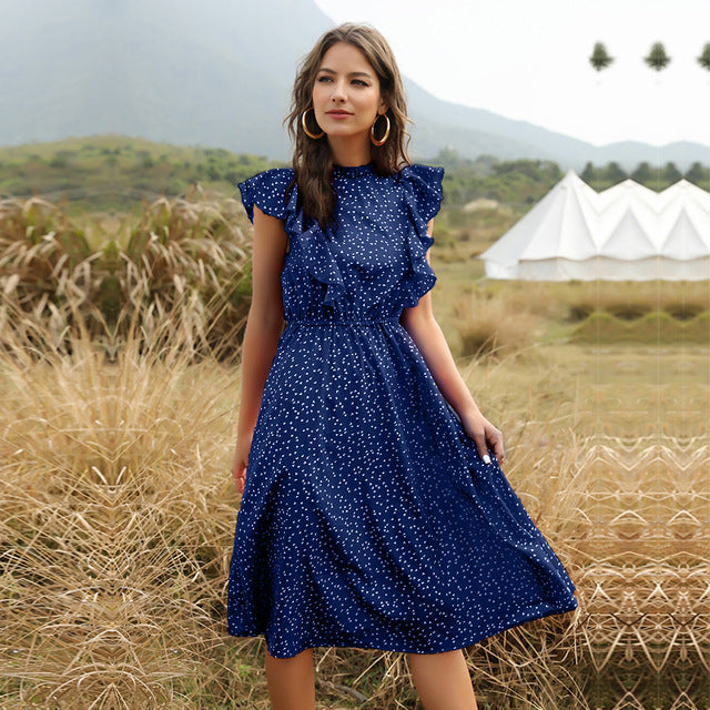 Frau in blauem gepunktetem Kleid steht in natürlicher Landschaft mit Bergen und Zelt im Hintergrund, Mode, Outdoor, Sommerkleid.