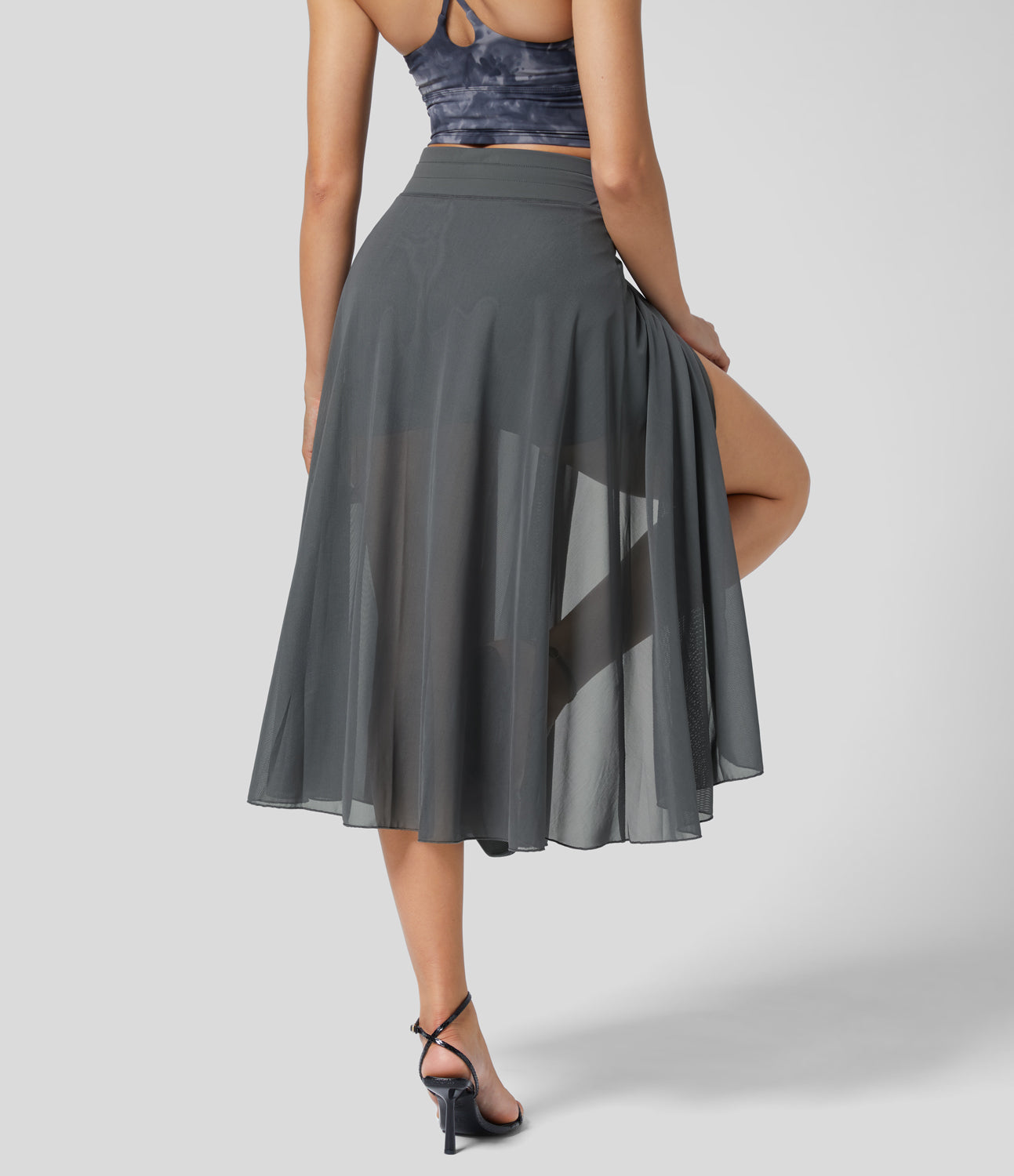Frau in grauem, transparentem Chiffon-Rock mit hohem Schlitz, kombiniert mit schwarzem Top und Absatzsandalen. Elegante Damenmode, Sommeroutfit.