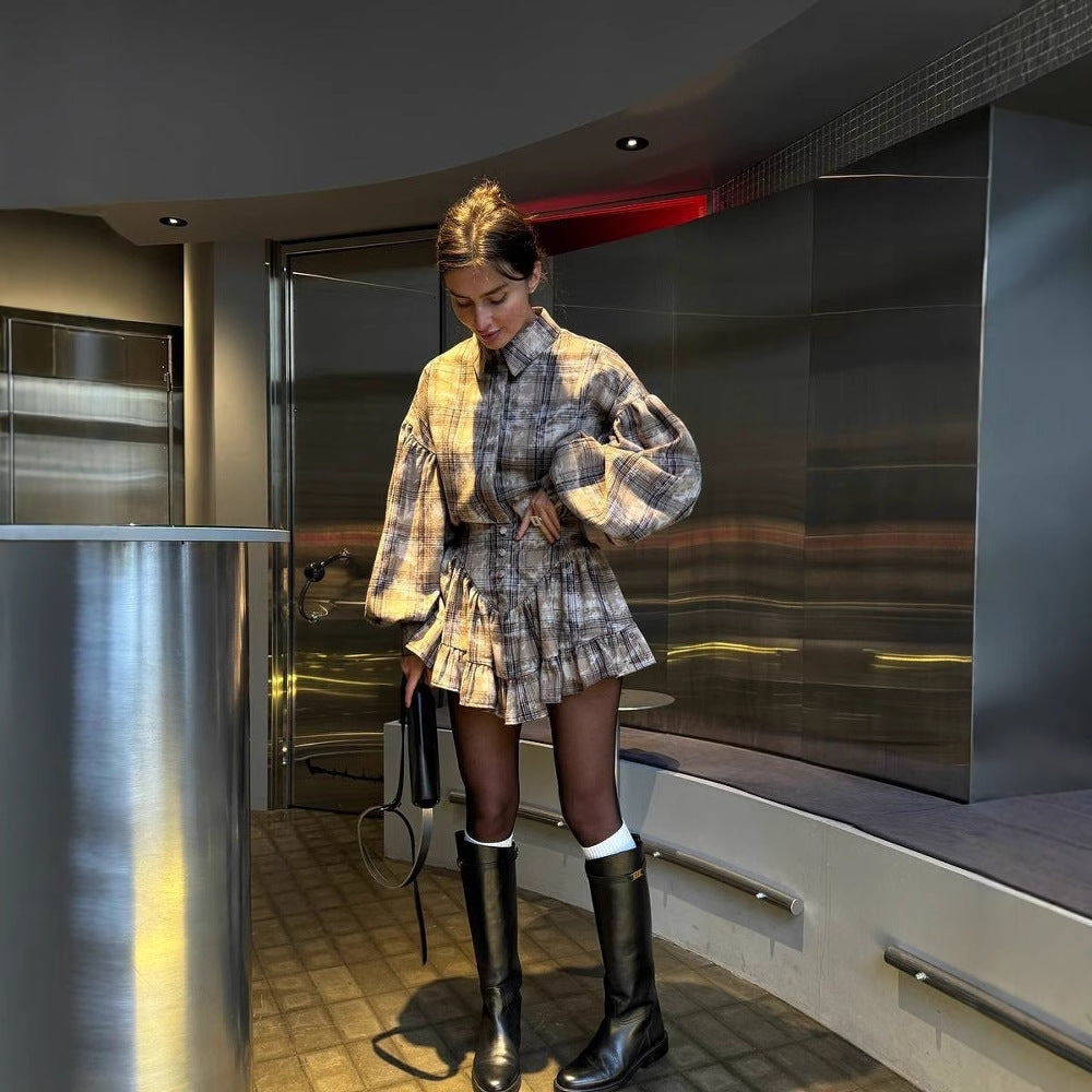 Frau in kariertem Kleid mit langen Ärmeln und schwarzen Stiefeln in modernem Innenraum. Modetrends, Damenmode, Herbstoutfit.