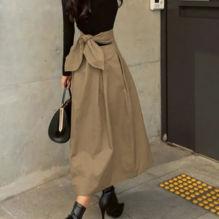 Frau in schwarzem Oberteil und beiger, knöchellanger Rock mit Schleife, schwarze Stiefeletten, schwarze Handtasche, moderne Mode, urbane Umgebung.