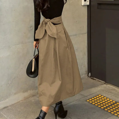 Frau in schwarzem Oberteil und beiger, knöchellanger Rock mit Schleife, schwarze Stiefeletten, schwarze Handtasche, moderne Mode, urbane Umgebung.