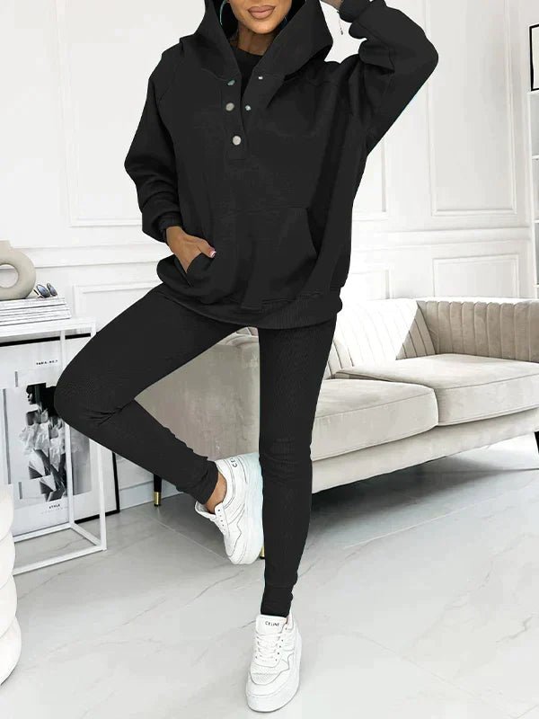 Dame in schwarzem Hoodie und Leggings posiert in modernem Wohnzimmer. Bequeme Freizeitmode, stilvoller Look, weiße Sneakers, minimalistische Einrichtung.