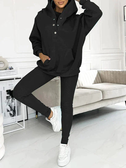 Dame in schwarzem Hoodie und Leggings posiert in modernem Wohnzimmer. Bequeme Freizeitmode, stilvoller Look, weiße Sneakers, minimalistische Einrichtung.