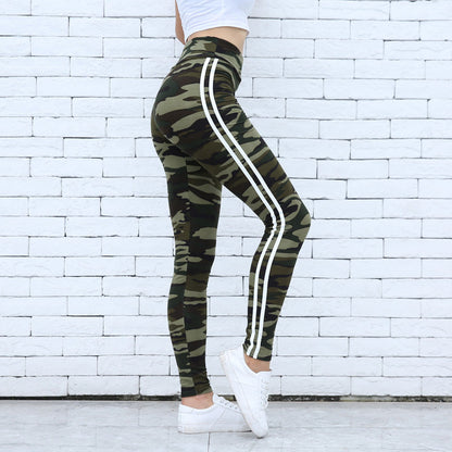 Frau in Camouflage-Leggings mit weißen Streifen, seitlich vor weißer Ziegelwand. Modetrend, Sportbekleidung, Streetwear-Look.