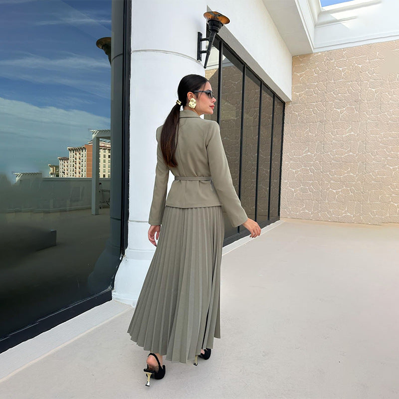 Frau in olivgrünem Blazer und plissiertem Rock, trägt Sonnenbrille und Ohrringe, geht auf modernem Balkon. Elegante Mode, stilvolle Kleidung.