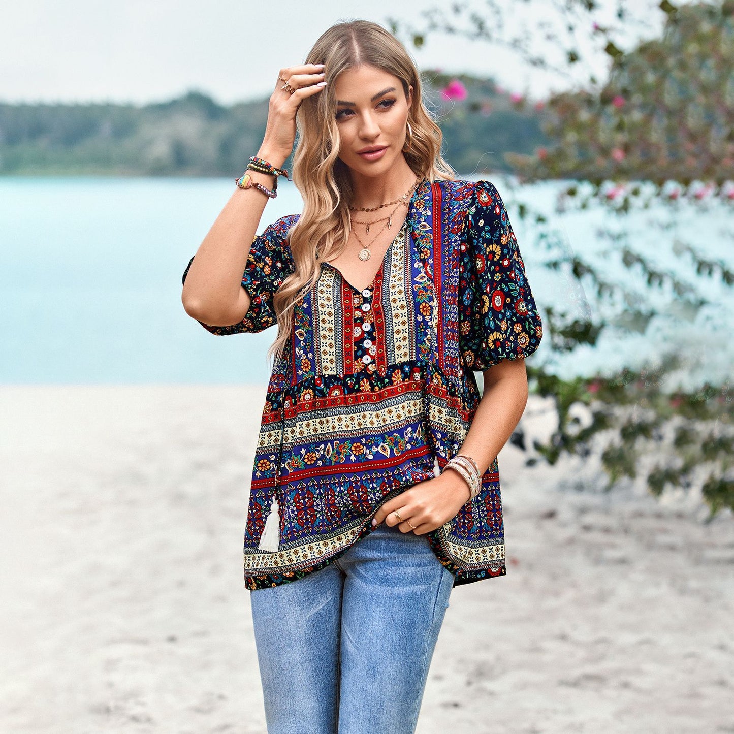 Frau in bunter Boho-Bluse und Jeans am Strand, umgeben von Natur. Sommermode, Freizeitkleidung, Strandoutfit, Boho-Stil.