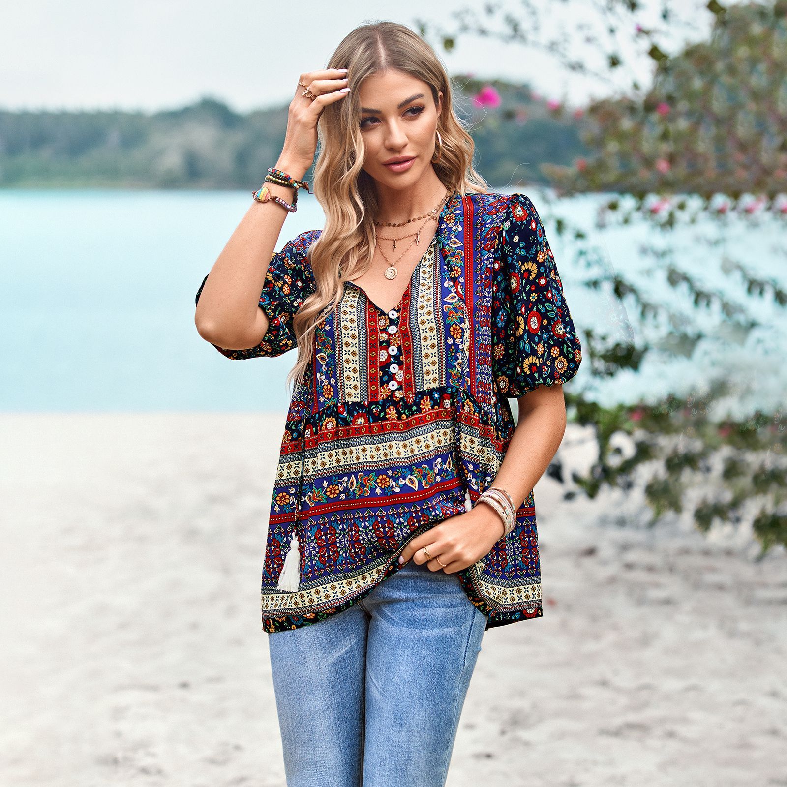 Frau in bunter Boho-Bluse und Jeans am Strand, umgeben von Natur. Sommermode, Freizeitkleidung, Strandoutfit, Boho-Stil.