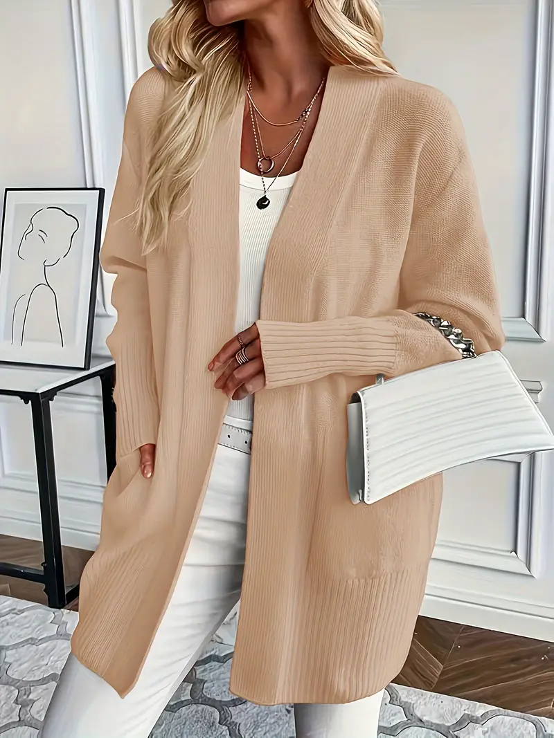 Blonde Frau in beigem Strickcardigan, weißem Top und Hose, hält weiße Handtasche. Elegante Mode, Herbstoutfit, stilvolle Damenbekleidung.