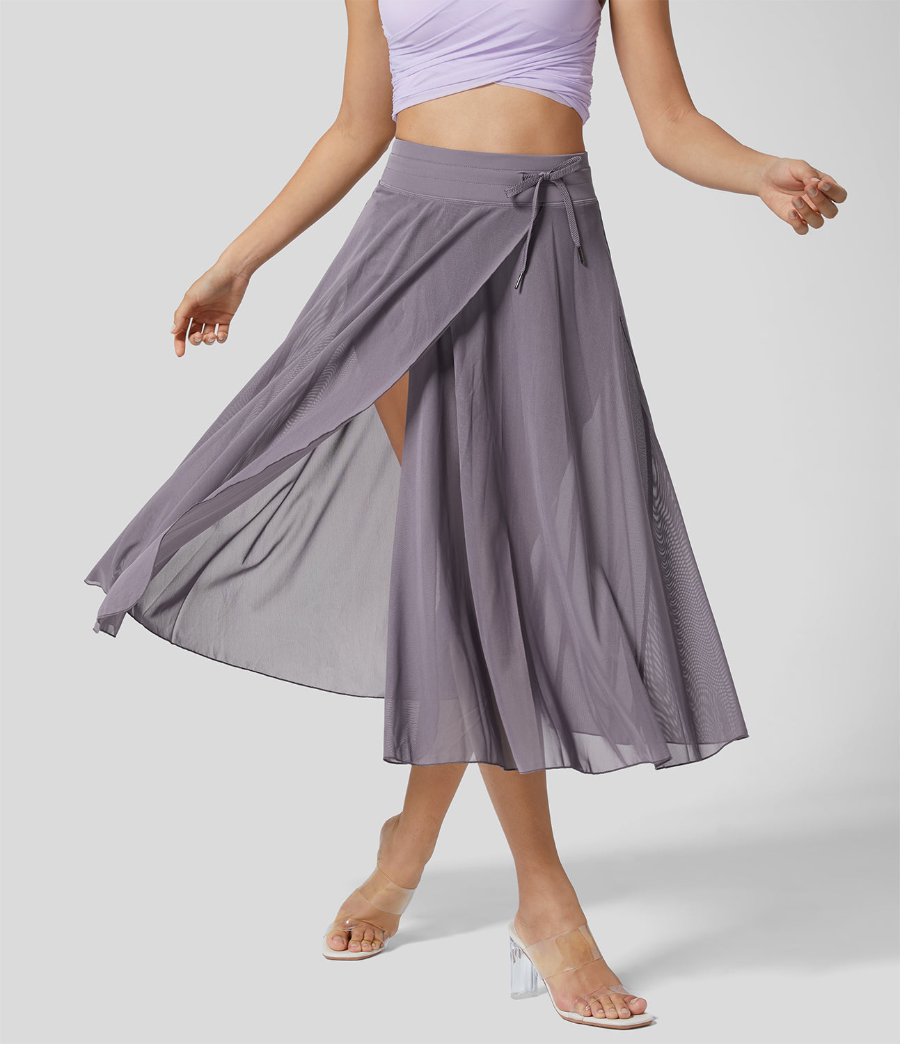 Frau trägt fliederfarbenen, fließenden Wickelrock mit Schlitz, kombiniert mit transparenten Absatzsandalen. Elegante Damenmode, Sommeroutfit.