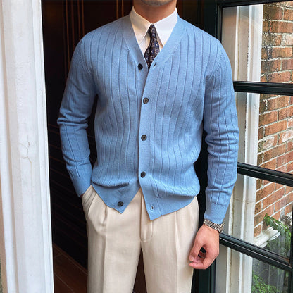 Mann in hellblauer Strickjacke mit Knöpfen, weißem Hemd und Krawatte, steht vor einem Fenster. Elegante Herrenmode, stilvoller Look.