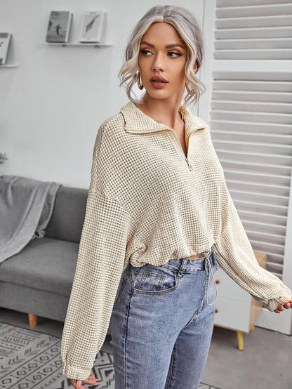 Blonde Frau in beigem Strickpullover und blauer Jeans steht in modernem Wohnzimmer. Stilvolle Mode, lässiger Look, Inneneinrichtung.