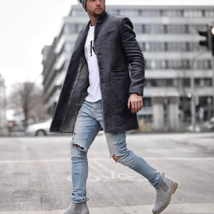 Mann in grauem Mantel, zerrissenen Jeans und grauen Stiefeln überquert Straße in urbaner Umgebung. Modischer Streetstyle, Wintermode.