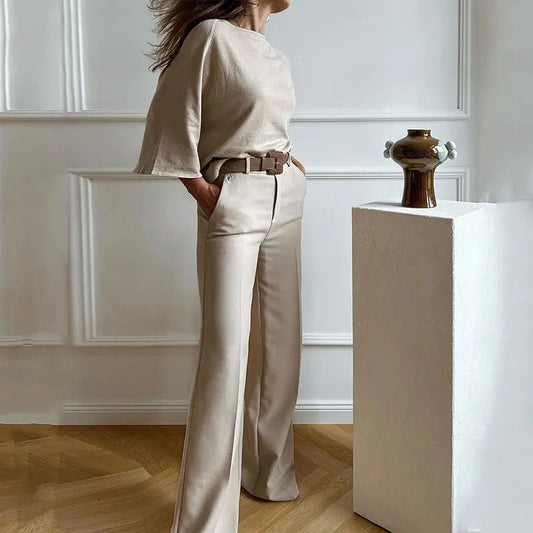 Elegante Frau in beiger Hose und Oberteil, steht neben einer modernen Vase auf einem Podest. Stilvolle Mode, minimalistische Inneneinrichtung.