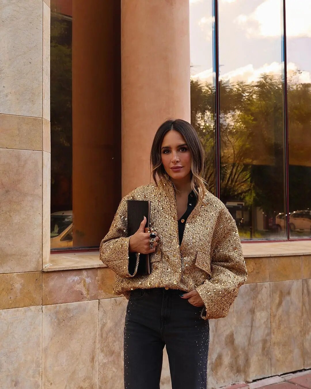 Frau in goldener Paillettenjacke und schwarzer Hose, hält schwarze Clutch, steht vor modernem Gebäude. Elegante Mode, Streetstyle, Herbstlook.
