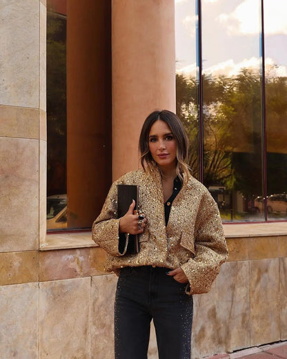 Frau in goldener Paillettenjacke und schwarzer Hose, hält schwarze Clutch, steht vor modernem Gebäude. Elegante Mode, Streetstyle, Herbstlook.
