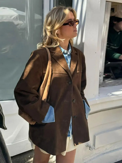 Frau in braunem Blazer und Sonnenbrille, trägt eine blaue Jeansjacke darunter, steht vor einem weißen Gebäude. Modetrend, Herbstmode, Streetstyle.