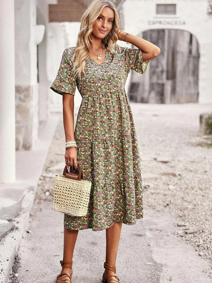 Frau in geblümtem Sommerkleid mit Strohtasche, steht auf gepflastertem Weg vor rustikalem Gebäude. Mode, Sommerkleid, Strohtasche, Outdoor.